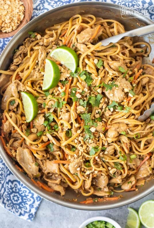 Skillet of Thai peanut noodles topped with chopped peanuts, lime wedges and chopped cilantro.