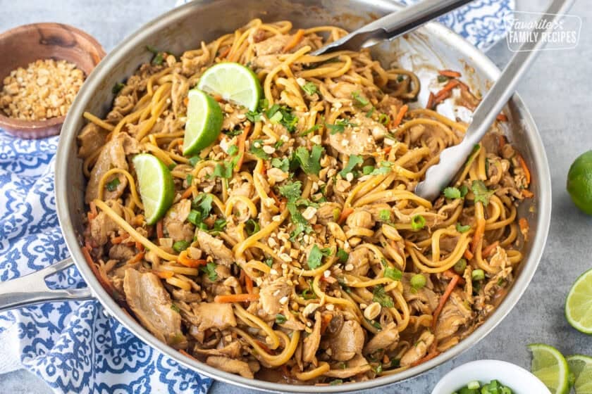 Skillet of Thai peanut noodles topped with chopped peanuts, lime wedges and chopped cilantro.