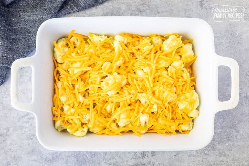 Baking dish of cauliflower mixed with sauce and shredded cheddar cheese on top.