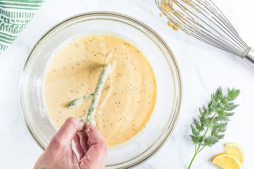 Two green beans being dipped into a clear bowl of a smooth batter