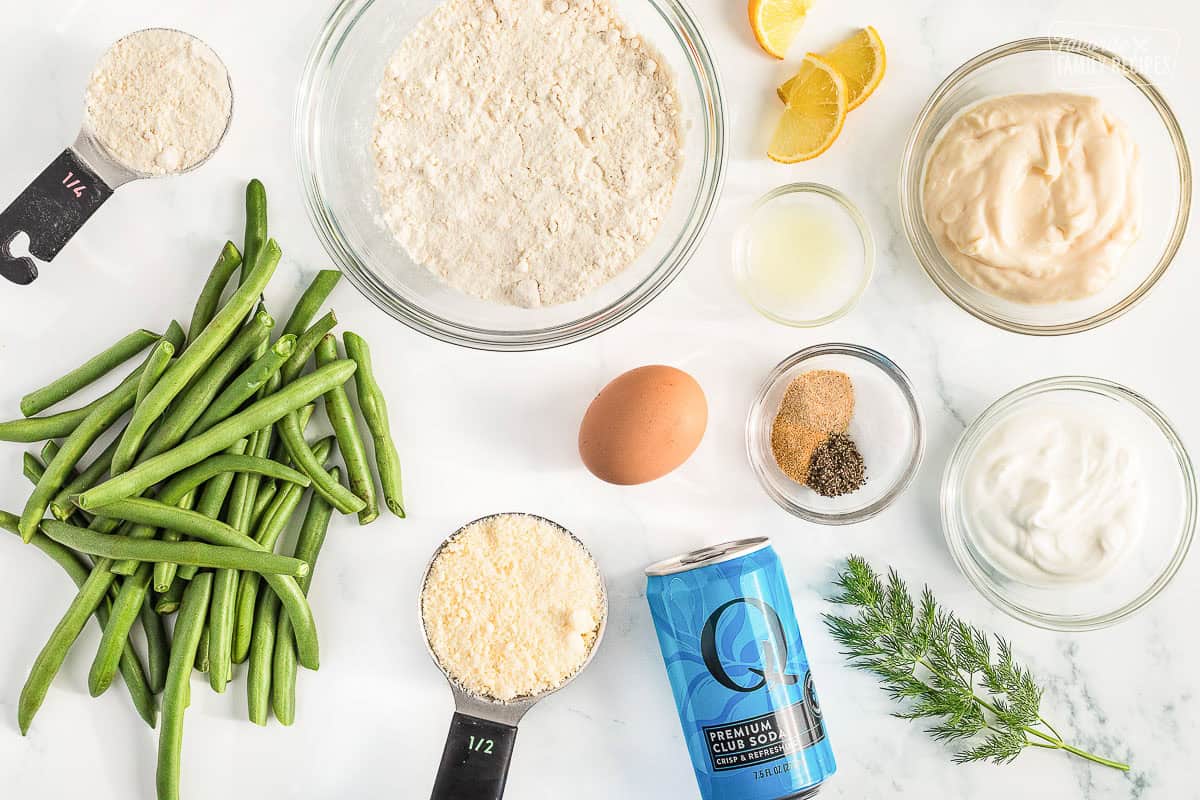 Ingredients to make fried green beans including club soda, flour, parmesan cheese, eggs, beans, lemon, dill, and seasonings laid out on a white counter top.
