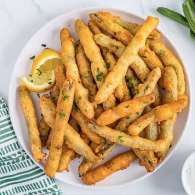 A top-down view of a plate of golden fried green beans garnished with two lemon wedges and a bowl of lemon dill dip in partial view on the side