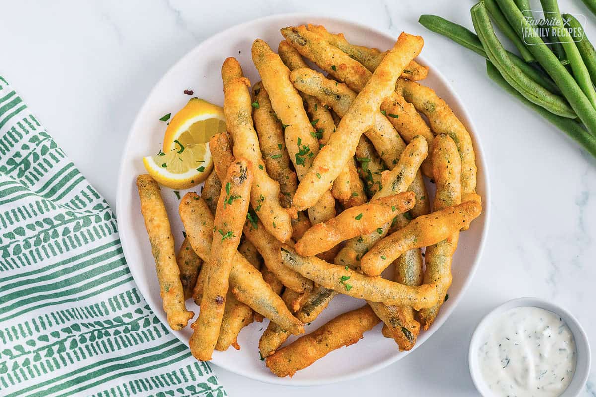 A top-down view of a plate of golden fried green beans garnished with two lemon wedges and a bowl of lemon dill dip in partial view on the side