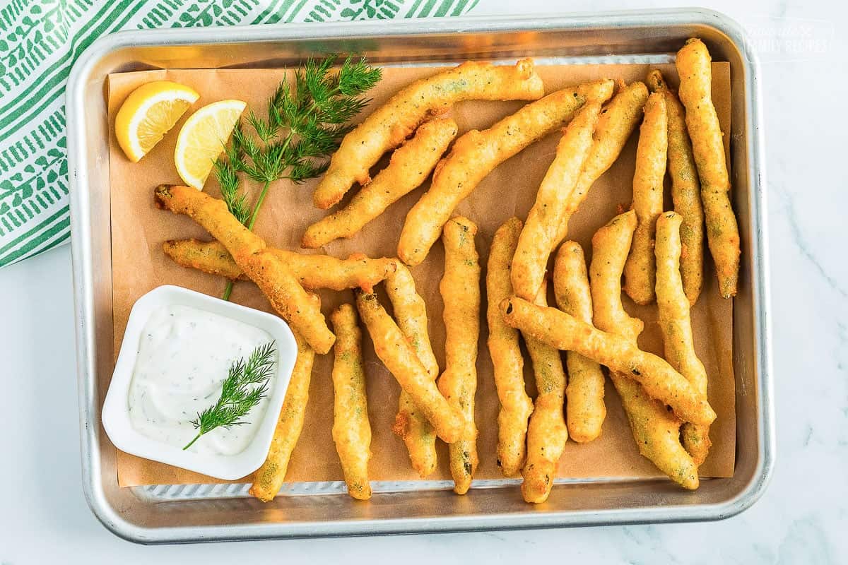 A rectangle tray lined with brown butcher paper topped with golden fried green beans and a small square bowl of lemon dill dip on the side
