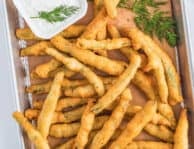 A rectangle tray lined with brown butcher paper topped with golden fried green beans and a small square bowl of lemon dill dip on the side