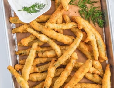 A rectangle tray lined with brown butcher paper topped with golden fried green beans and a small square bowl of lemon dill dip on the side