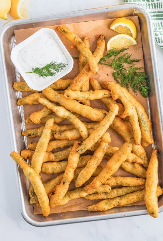 A rectangle tray lined with brown butcher paper topped with golden fried green beans and a small square bowl of lemon dill dip on the side