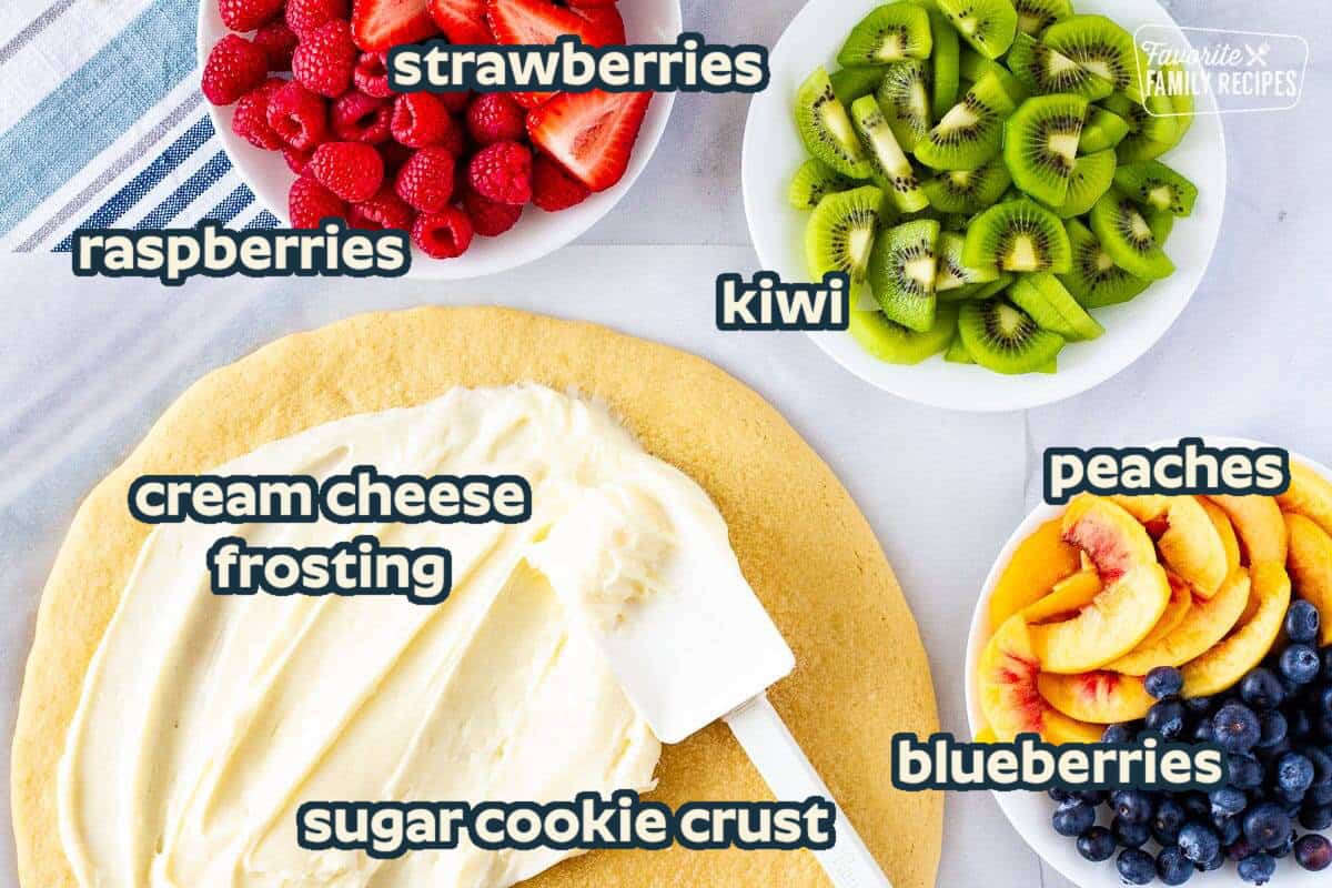 Cookie pizza with a spatula spreading cream cheese frosting and three bowls of fresh fruit on the side. Fruits include raspberries, strawberries, kiwi, peaches, and blueberries.