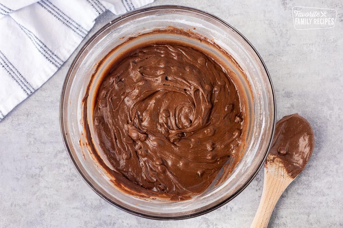 Homemade brownie batter in a glass mixing bowl and wooden spoon on the side.