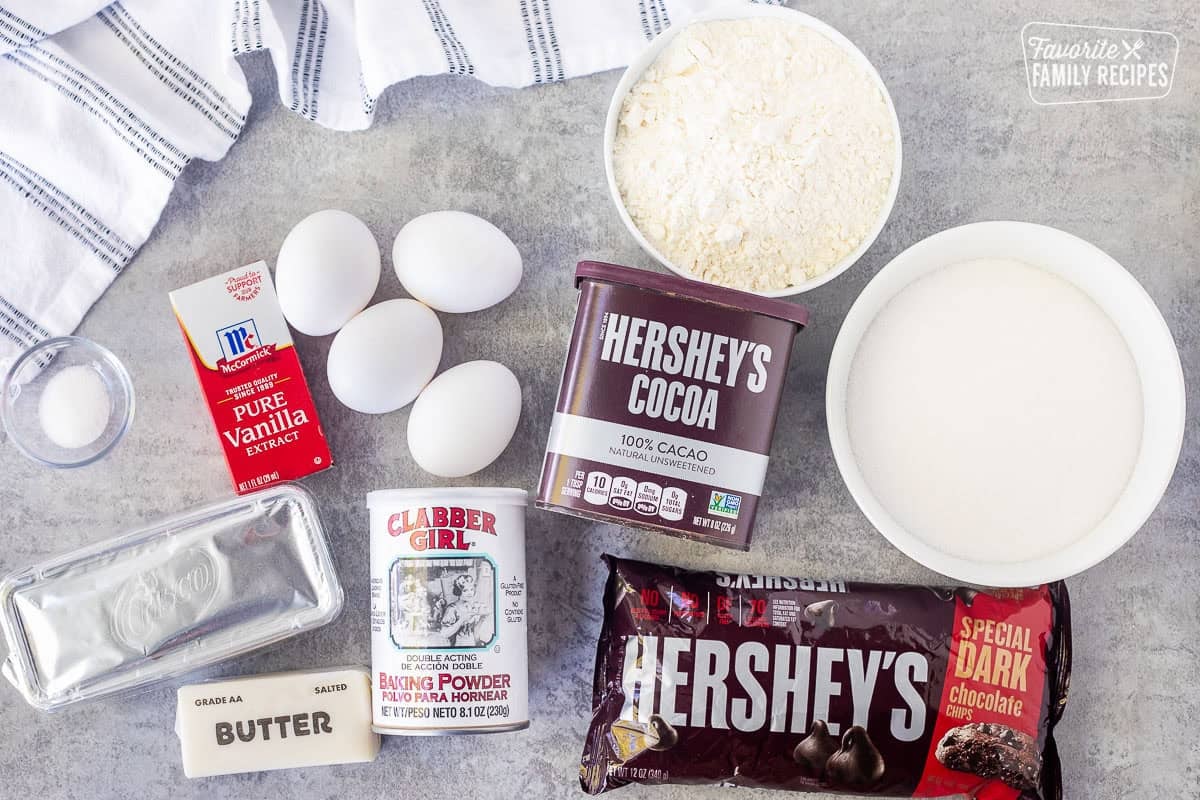 Ingredients to make homemade brownies including cocoa, flour, sugar, dark chocolate chips, baking powder, butter, shortening, vanilla, eggs and salt.