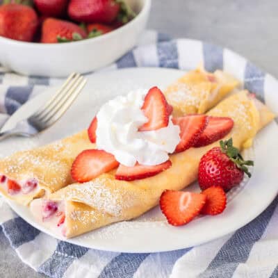 Two strawberry crepes on a plate with sliced strawberries and whipped cream.