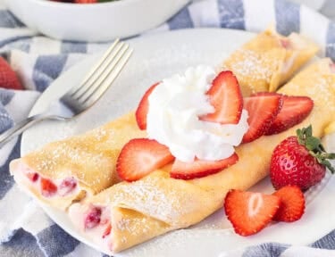 Two strawberry crepes on a plate with sliced strawberries and whipped cream.