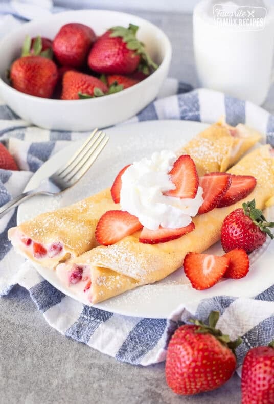 Two strawberry crepes on a plate with sliced strawberries and whipped cream.