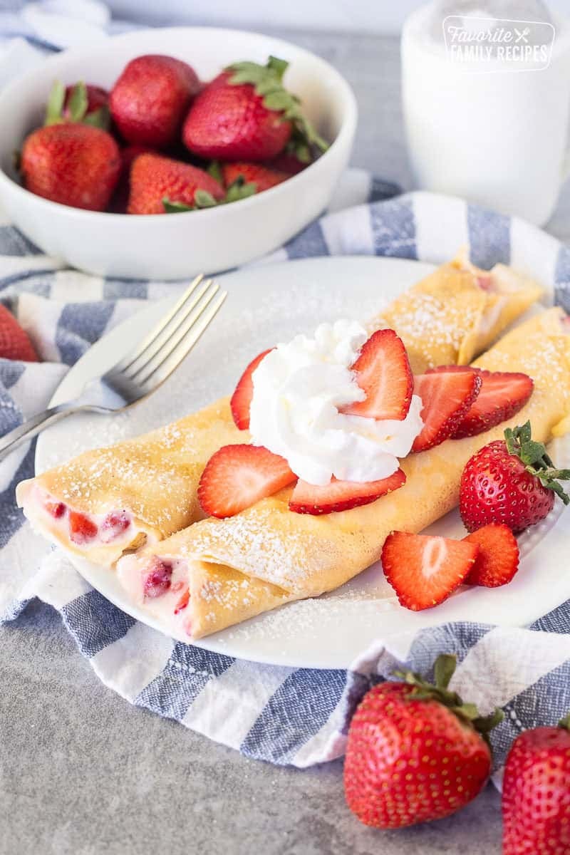 Two strawberry crepes on a plate with sliced strawberries and whipped cream.