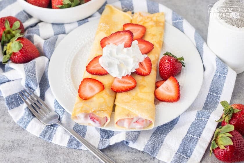 Two strawberry crepes on a plate with sliced strawberries and whipped cream.