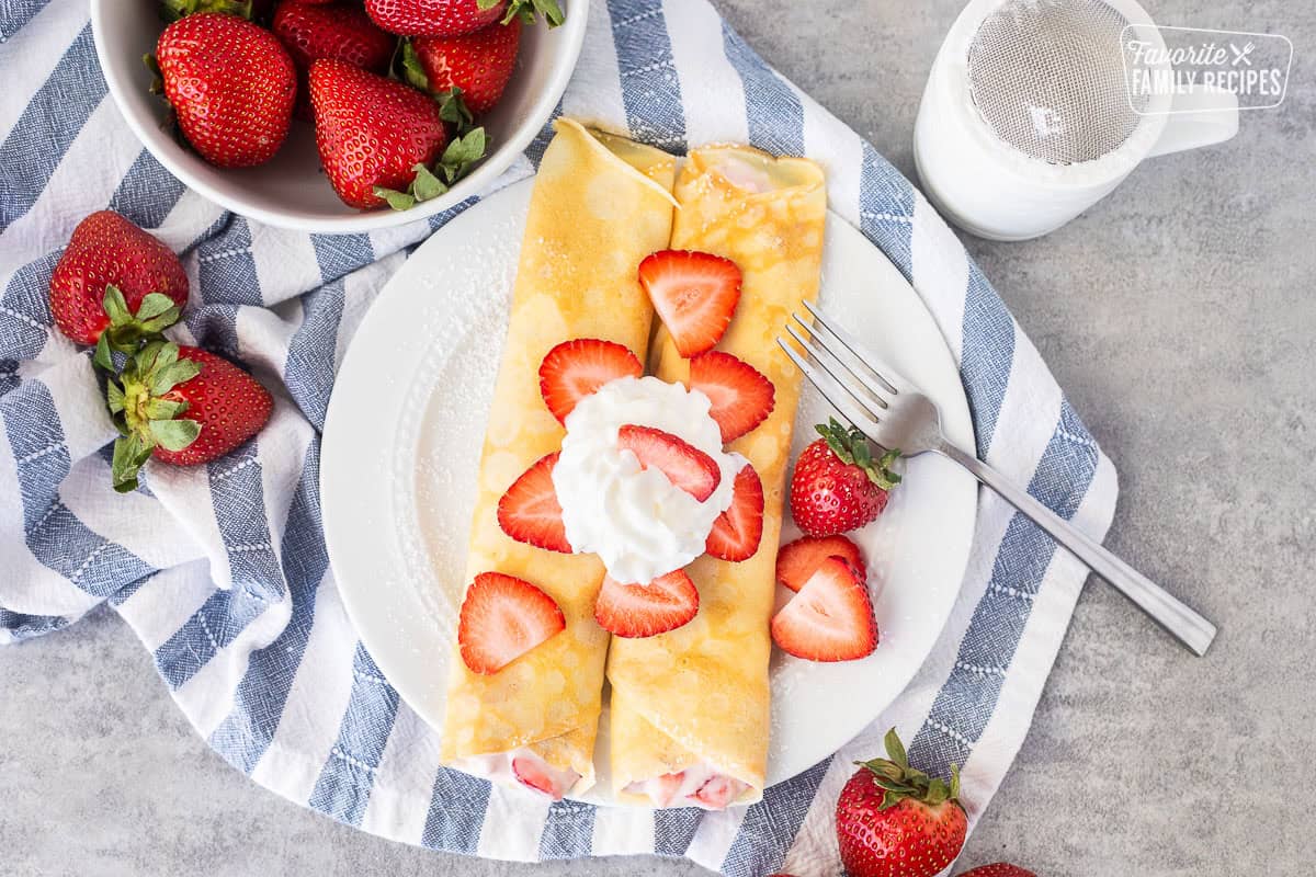 Two strawberry crepes on a plate with sliced strawberries and whipped cream.