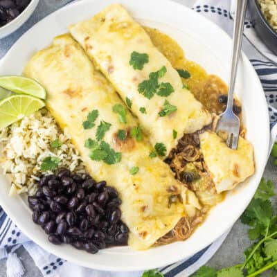 Angled view of two sweet pork enchiladas with green enchilada sauce on a white plate with cilantro lime rice, black beans, and lime wedges on the side with a fork cutting into one of the enchiladas.