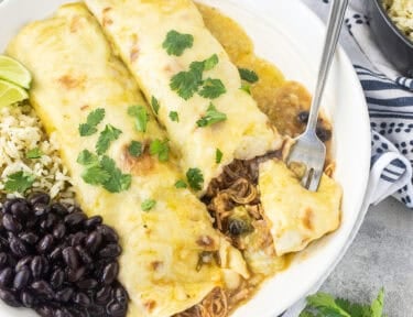Angled view of two sweet pork enchiladas with green enchilada sauce on a white plate with cilantro lime rice, black beans, and lime wedges on the side with a fork cutting into one of the enchiladas.