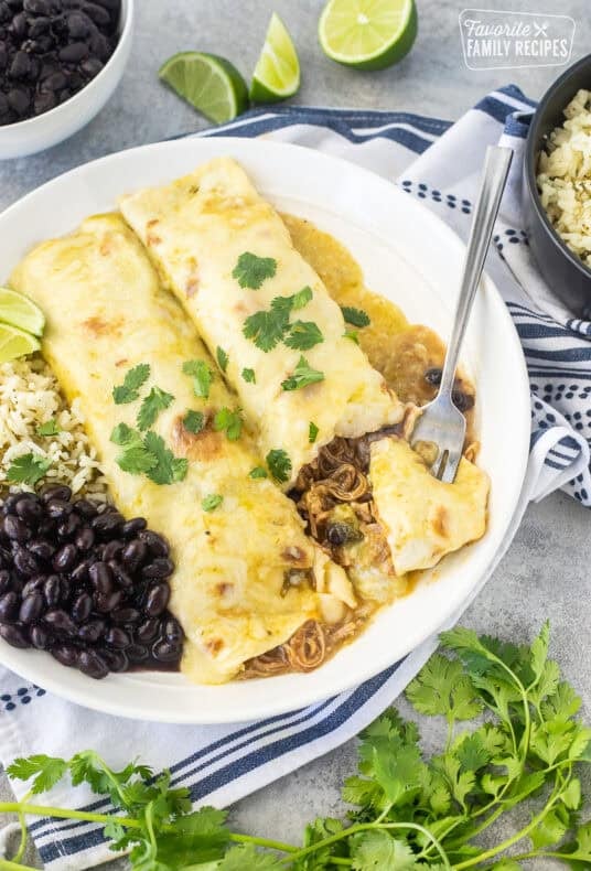 Angled view of two sweet pork enchiladas with green enchilada sauce on a white plate with cilantro lime rice, black beans, and lime wedges on the side with a fork cutting into one of the enchiladas.
