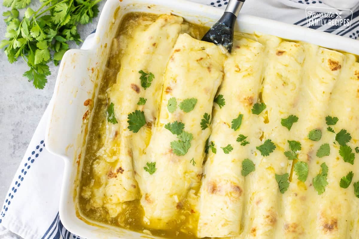 Spatula scooping up a sweet pork enchilada with green enchilada sauce from a white baking dish to show cheesy texture.