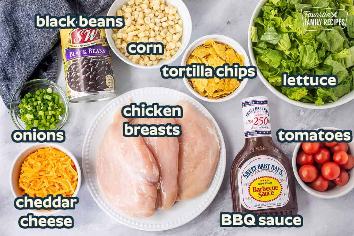 Ingredients to make BBQ Chicken Salad including chicken breasts, barbecue sauce, tomatoes, lettuce, chips, corn, black beans, green onions, and cheddar cheese.