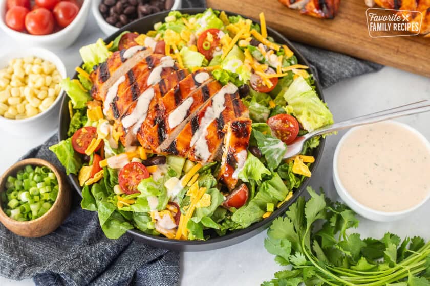 Fork with piece barbecue grilled chicken salad topped with black beans, corn, tomatoes, crushed tortilla chips, green onion and shredded cheddar cheese. Drizzled with creamy barbecue dressing.