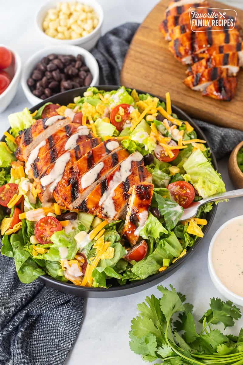 Fork with piece barbecue grilled chicken salad topped with black beans, corn, tomatoes, crushed tortilla chips, green onion and shredded cheddar cheese. Drizzled with creamy barbecue dressing.