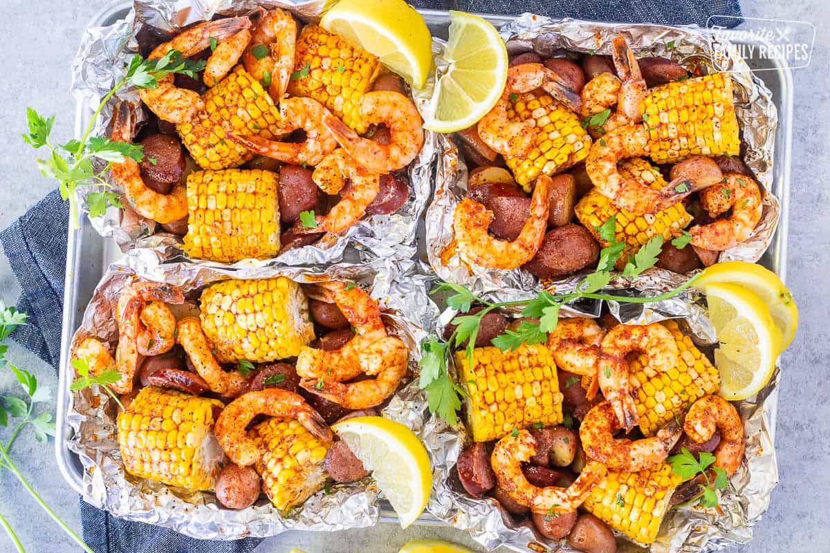 Four Cajun Foil Packets with shrimp, sausage, corn and red potatoes. Garnished with parsley and lemon wedges.