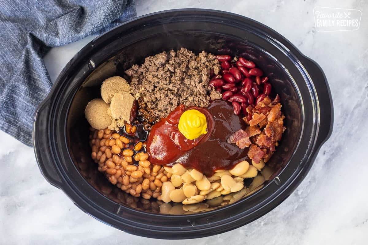 Crockpot of calico beans ingredients including ground beef, pork and beans, onion, garlic, ketchup, barbecue sauce, butter beans, kidney beans, bacon, brown sugar, salt, pepper, molasses, and mustard.