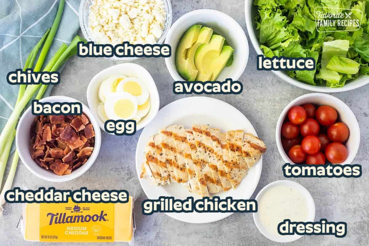 Ingredients to make Cobb salad including grilled chicken breast, tomatoes, lettuce, avocado, cheddar cheese, green onions, hard boiled eggs, crumbled blue cheese, bacon and ranch dressing.