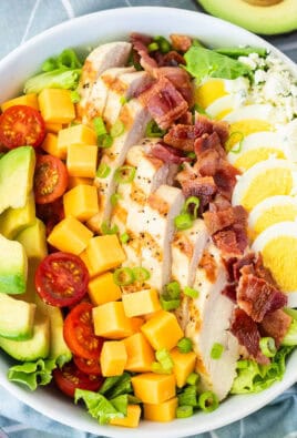 Cobb salad in a bowl with lettuce, blue cheese, sliced hard boiled egg, bacon, grilled chicken, cubed cheddar cheese, tomatoes and sliced avocado. Topped with green onion and Ranch dressing on the side.
