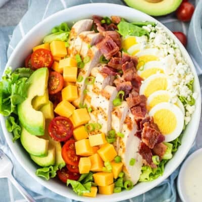 Cobb salad in a bowl with lettuce, blue cheese, sliced hard boiled egg, bacon, grilled chicken, cubed cheddar cheese, tomatoes and sliced avocado. Topped with green onion and Ranch dressing on the side.