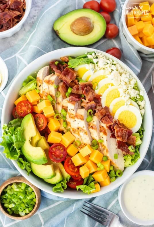 Cobb salad in a bowl with lettuce, blue cheese, sliced hard boiled egg, bacon, grilled chicken, cubed cheddar cheese, tomatoes and sliced avocado. Topped with green onion and Ranch dressing on the side.