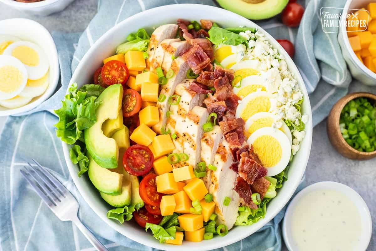 Cobb salad in a bowl with lettuce, blue cheese, sliced hard boiled egg, bacon, grilled chicken, cubed cheddar cheese, tomatoes and sliced avocado. Topped with green onion and Ranch dressing on the side.