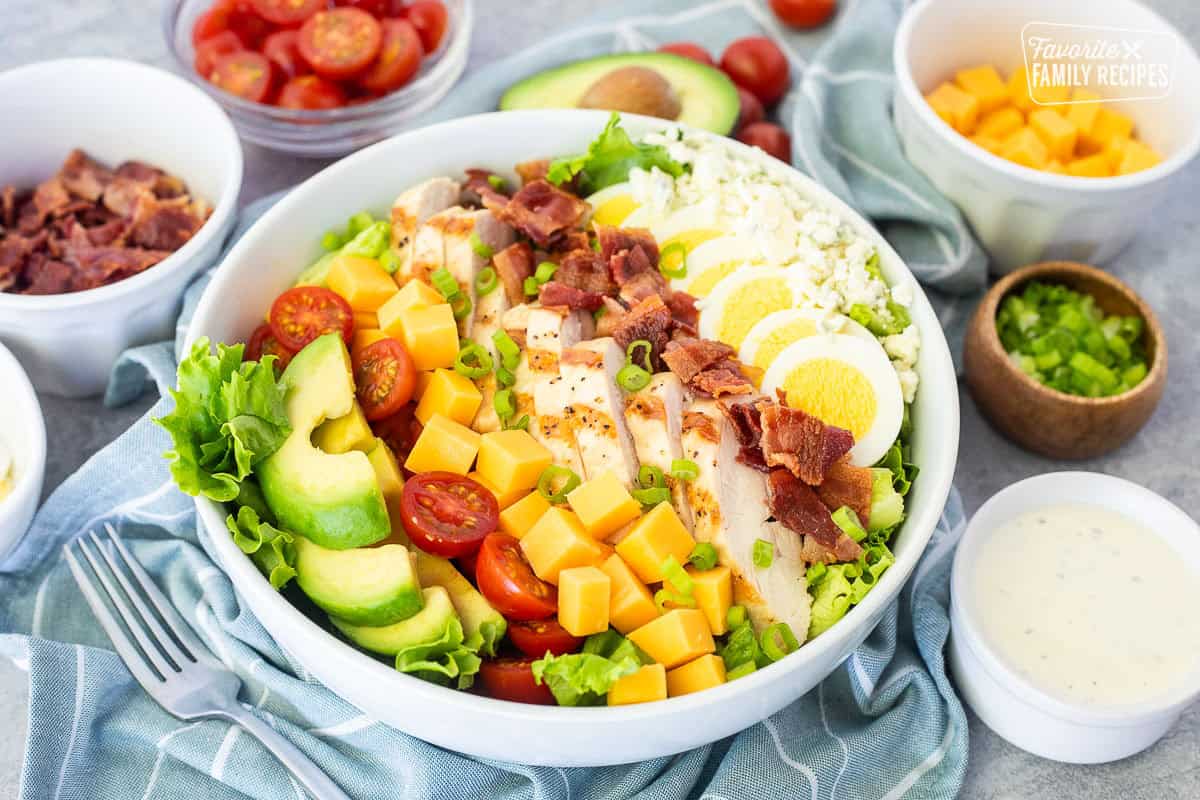 Cobb salad in a bowl with lettuce, blue cheese, sliced hard boiled egg, bacon, grilled chicken, cubed cheddar cheese, tomatoes and sliced avocado. Topped with green onion and Ranch dressing on the side.