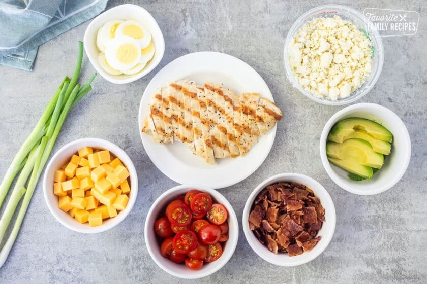 Toppings for Cobb salad including grilled chicken breast, sliced cherry tomatoes, cut avocado, cubed cheddar cheese, green onions, hard boiled eggs, crumbled blue cheese, and bacon pieces.