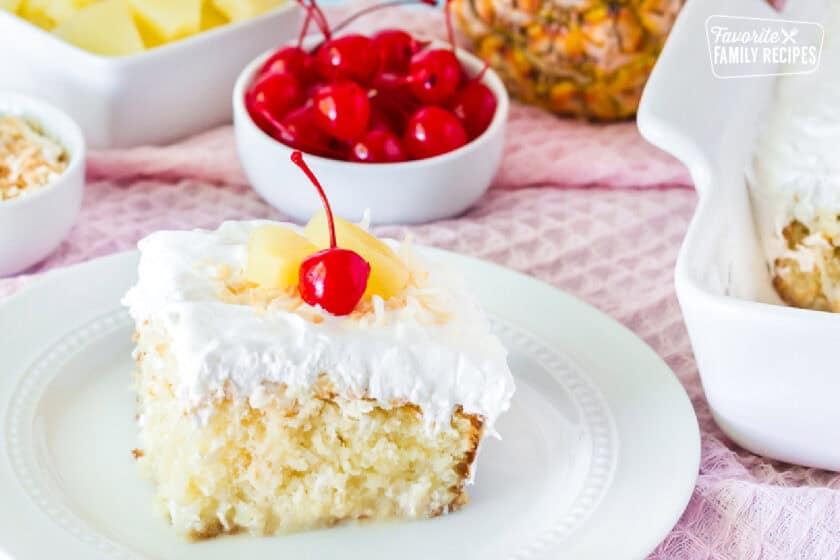Slice of Pina Colada Cake on a plate. Fresh pineapple, cherries, toasted coconut and pineapple slices on the side.
