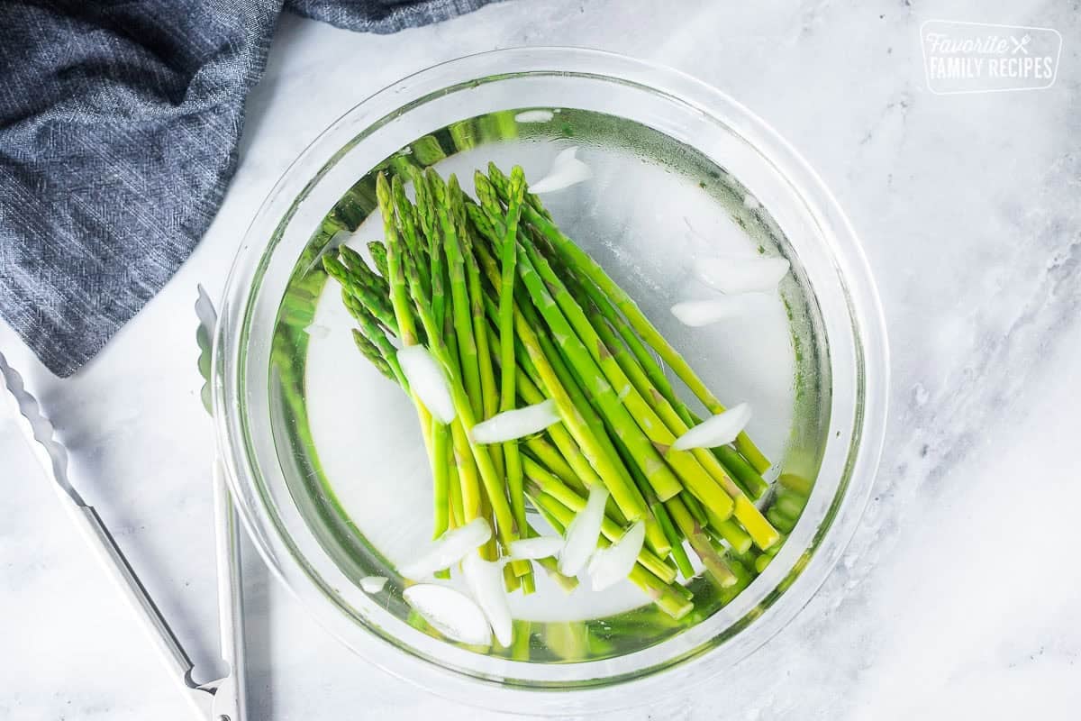 Glass bowl with ice water and asparagus. Tongs on the side.