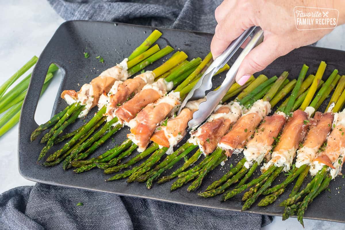 Picking up prosciutto wrapped asparagus with tongs from tray.
