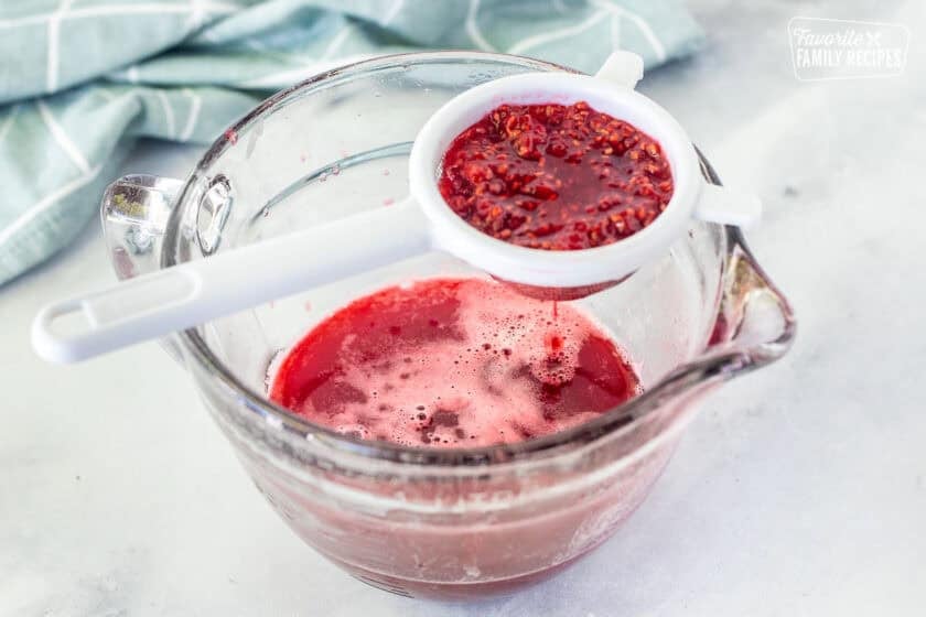 Straining raspberries with mesh strainer over a glass pouring bowl.