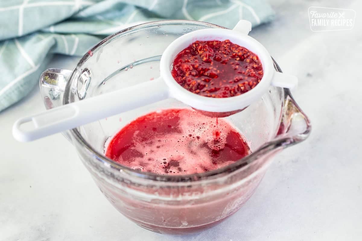 Straining raspberries with mesh strainer over a glass pouring bowl.