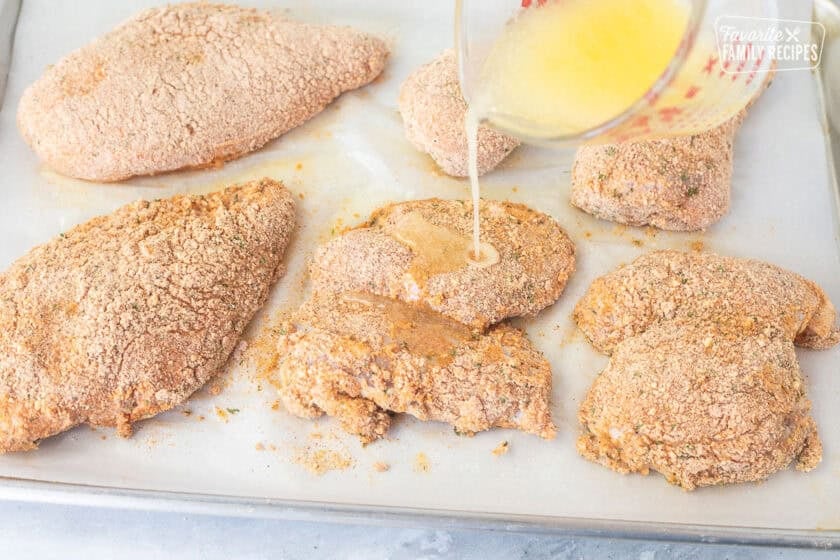 Pouring butter oil mixture on top of coated chicken pieces.