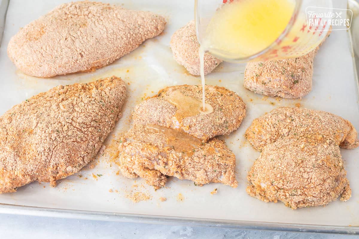Pouring butter oil mixture on top of coated chicken pieces.