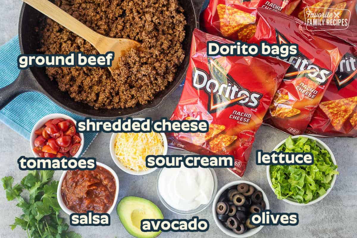 Ingredients to make walking tacos including Doritos, lettuce, black olives, sour cream, avocado, shredded cheese, salsa, tomatoes and seasoned ground beef.