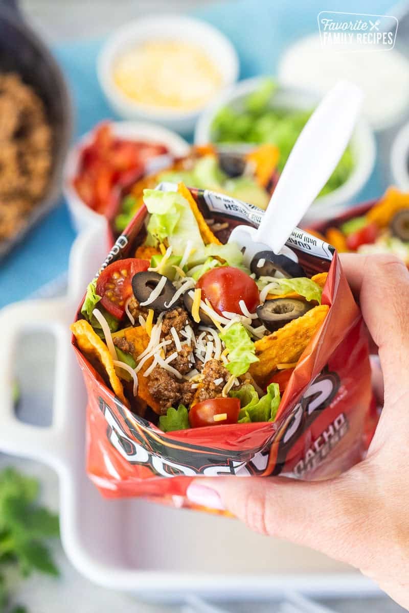 Hand holding a walking taco with nacho cheese Doritos, seasoned ground beef, cheese, lettuce, tomatoes, olives, avocado and a fork.