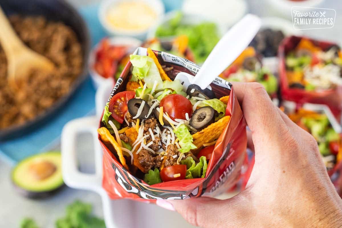 Hand holding a walking taco with nacho cheese Doritos, seasoned ground beef, cheese, lettuce, tomatoes, olives, avocado and a fork.