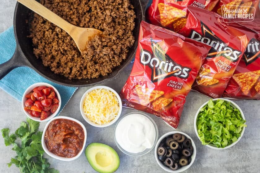 Ingredients to make walking tacos including Doritos, lettuce, black olives, sour cream, avocado, shredded cheese, salsa, tomatoes and seasoned ground beef.