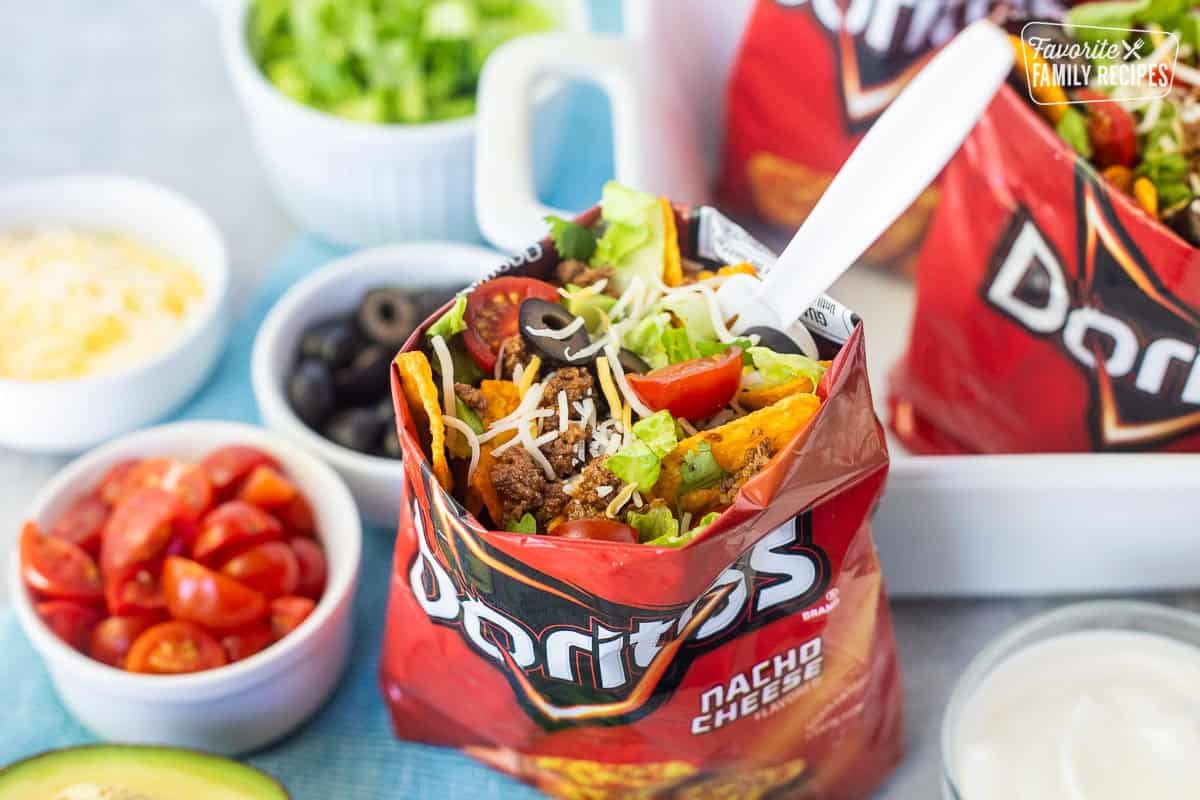 Walking taco with nacho cheese Doritos, seasoned ground beef, cheese, lettuce, tomatoes, olives, avocado and a fork.