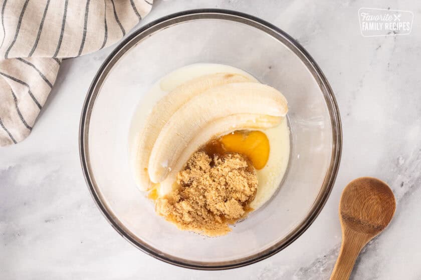 Glass mixing bowl with ripe peeled bananas, brown sugar, egg, vanilla, oil and milk. Wooden spoon on the side.