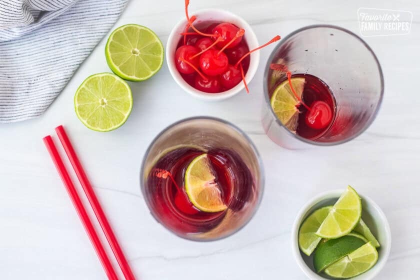 Two glasses with a lime wedge, cherry and cherry syrup on the bottom.
